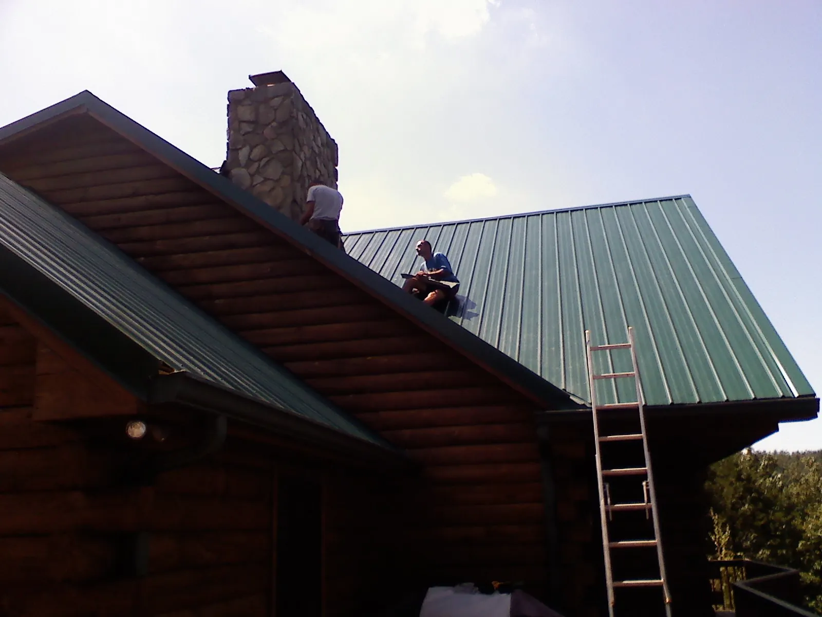 Roofers installing green metal panels beside a stone chimney on a log home in New Park